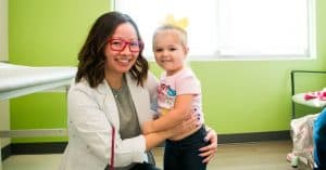 A Pediatrician from Pediatric Associates of Dallas smiling beside a young girl in a medical exam room