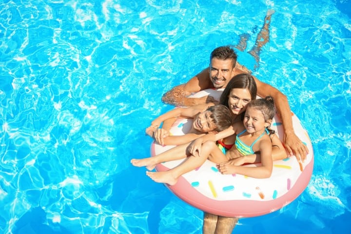 Happy family of four, two adults and two children, floating on a large pink doughnut-shaped pool ring in a bright blue swimming pool, enjoying a sunny day