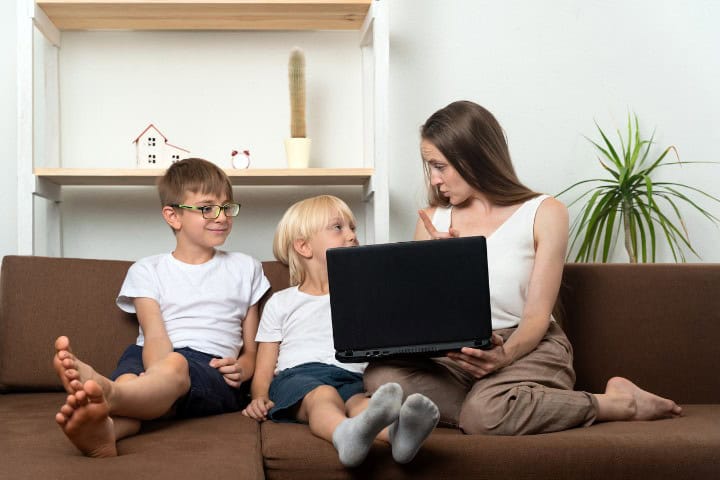 A mother explaining something to her two young children while they look at a laptop on a couch