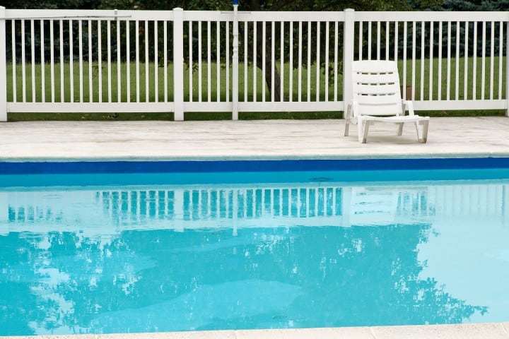 serene poolside scene with a single white lounge chair beside calm blue water, reflecting a white fence and lush greenery