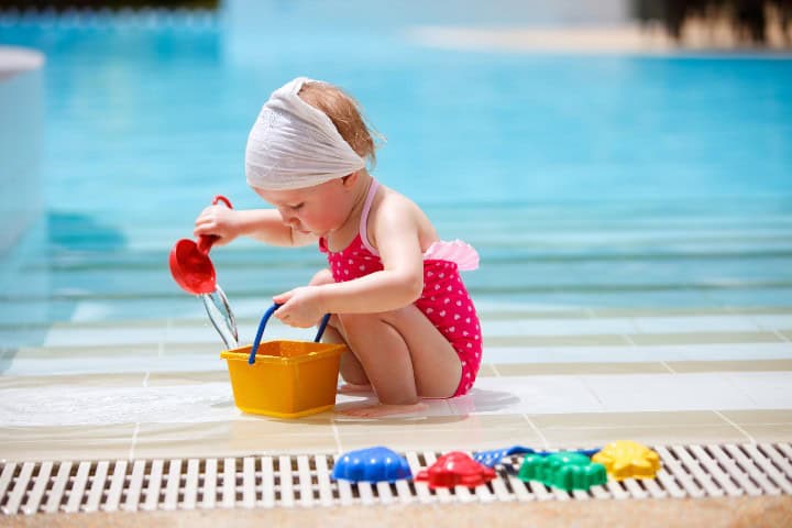 Young toddler in a pink polka-dotted swimsuit and white head wrap, playing with a yellow bucket and red shovel at the pool edge