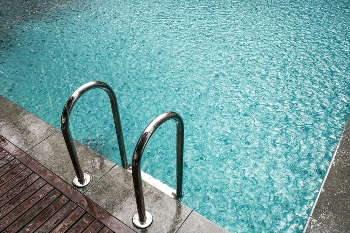 Stainless steel ladder leading into a turquoise swimming pool, with raindrops creating a shimmering effect on the water's surface