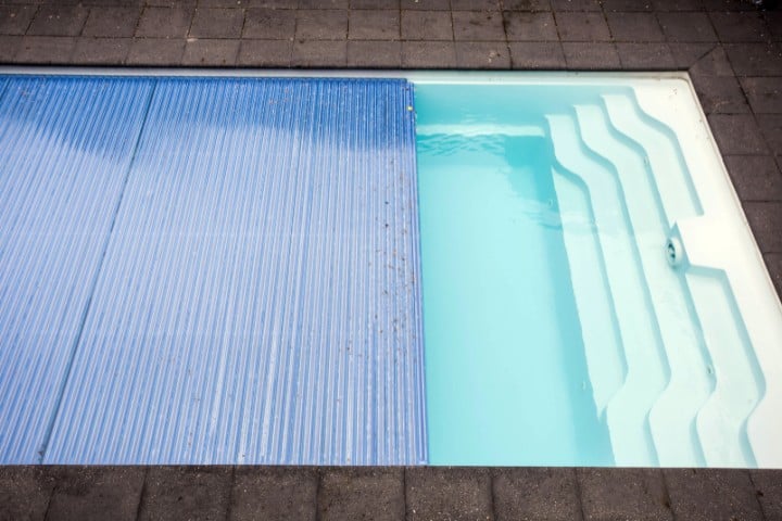 Partial view of a swimming pool with a sturdy blue cover partially retracted to reveal clear turquoise water underneath