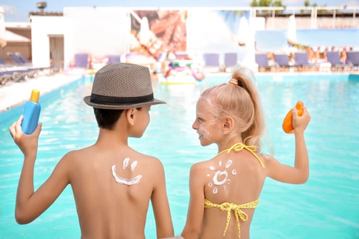 Two children by a poolside, creatively applying sunscreen; the boy with a handprint and the girl with playful designs on their backs