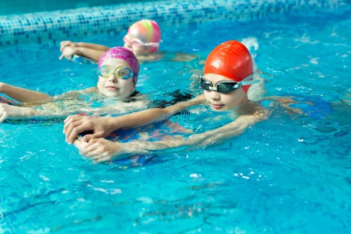 Three children in a swimming pool, each wearing colorful swim caps and goggles, using a floatation device for support