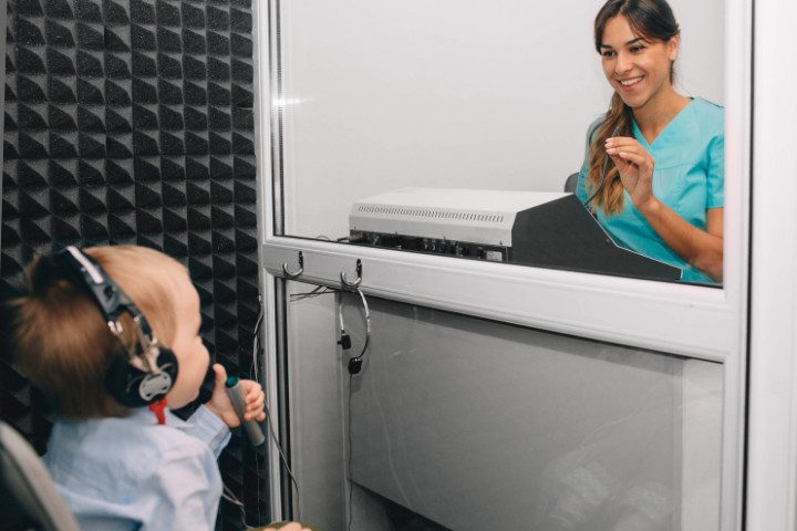 An audiologist conducting a hearing test for a young boy wearing headphones in a soundproof room