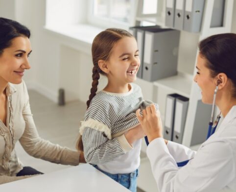 A smiling pediatrician examining a young girl with a stethoscope while the girl's mother watches in a clinic