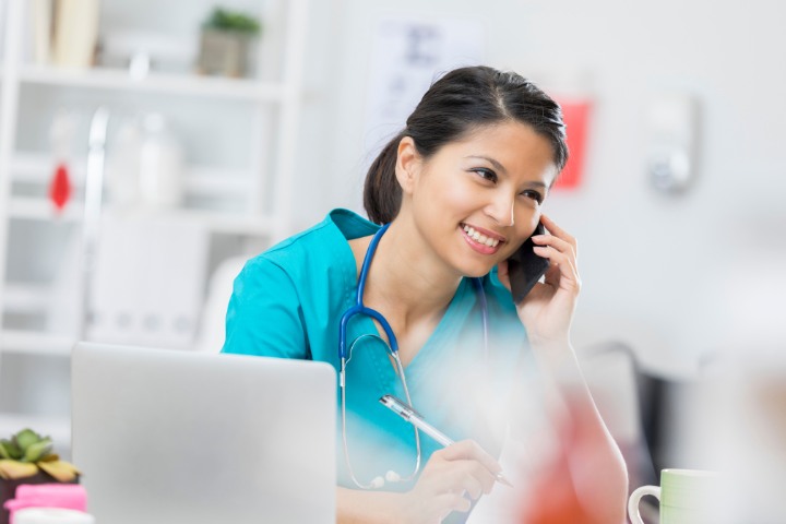 Smiling nurse in blue scrubs on the phone while working at her desk with a laptop