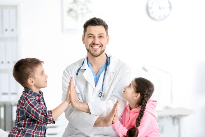 A smiling pediatrician giving high-fives to two young children in a clinic setting