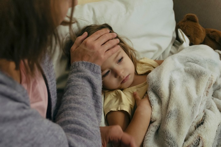 A sick child resting under a blanket while being comforted by an adult checking her temperature