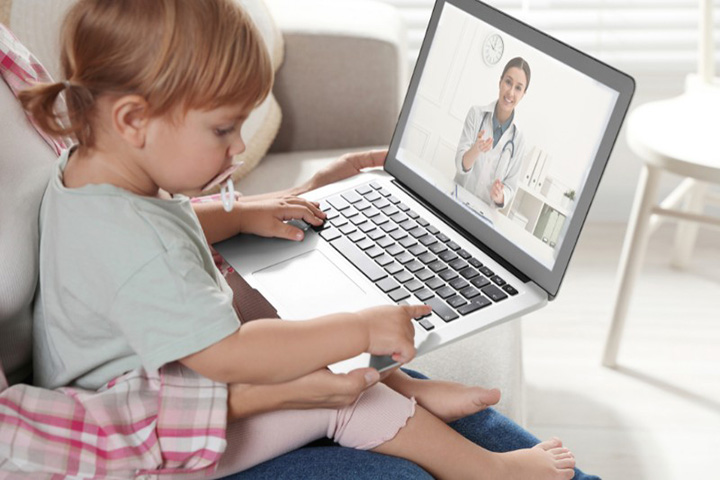 A toddler sitting on an adult's lap during a virtual consultation with a pediatrician displayed on a laptop